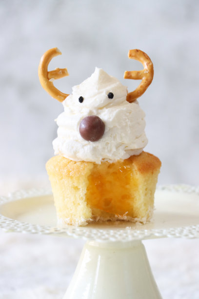 个人的特写图片，自制的圣诞驯鹿设计纸杯蛋糕在蛋糕架上，缺失的咬痕显示杏果酱馅，奶油漩涡顶部装饰着糖果包裹的巧克力甜鼻子和咸椒盐脆饼鹿角，重点在前景图片下载