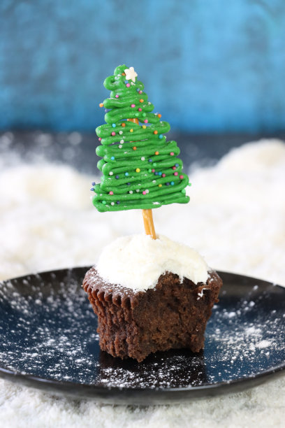 个人的特写图片，自制的，圣诞树设计的巧克力纸杯蛋糕在一个盘子里，奶油涂在干燥的椰子浇头，白巧克力和椒盐脆饼棒树，糖粉雪，蓝色背景，重点在前景图片下载