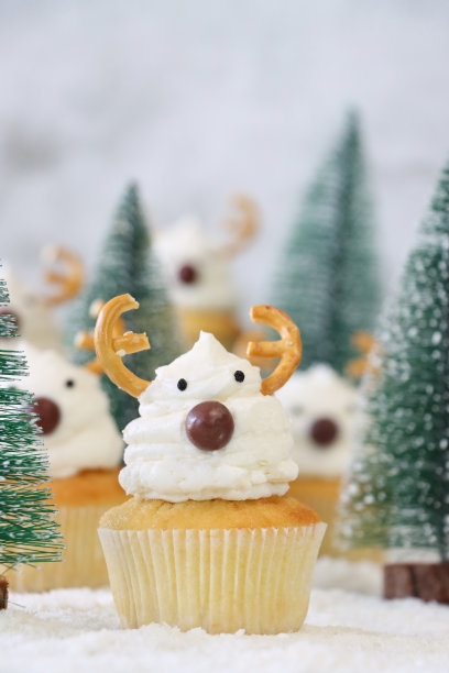 这批自制的圣诞驯鹿设计纸杯蛋糕的特写图片，奶油漩涡顶部装饰着糖果包裹的巧克力甜鼻子和咸椒盐卷饼鹿角，展示在白雪皑皑的针叶林场景中，重点放在前景图片下载