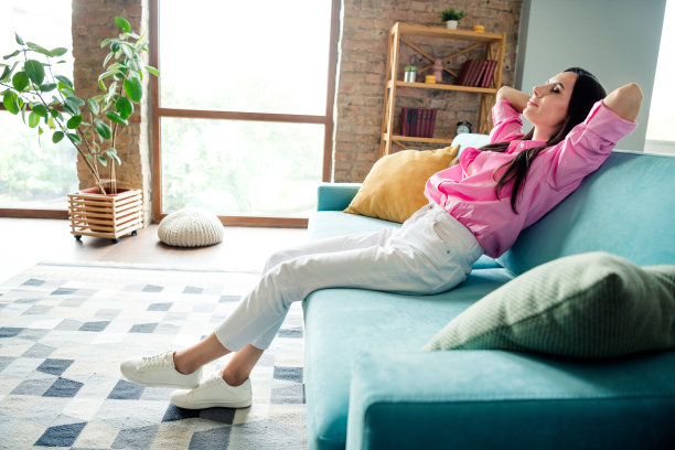 照片中，心情好，积极向上的年轻女子穿着粉色衬衫，坐在沙发上，双手抱在脑后，闭着眼睛，在公寓房间里图片下载