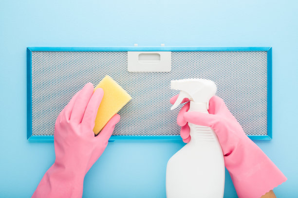 妇女双手戴上橡胶保护手套，手里拿着海绵和洗涤剂瓶，正在清洁厨房灶具罩过滤器，背景是蓝色的桌子。柔和的颜色。特写镜头。视角镜头。自顶向下视图。图片下载