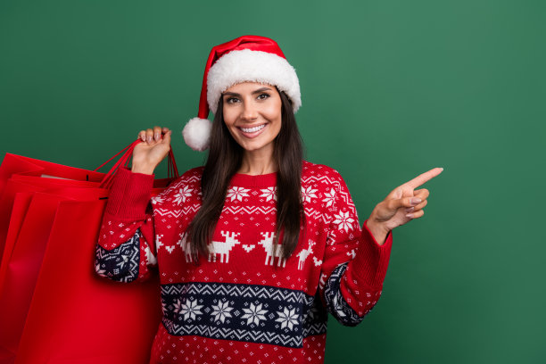 照片中开朗甜美的圣诞老人帮手女士穿着印花圣诞毛衣抱着购物者空空荡荡的绿色背景图片下载