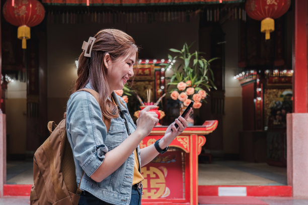 身穿蓝色夹克的年轻亚洲女子用相机或手机在中国寺庙拍摄欢快的照片或自拍。图片下载