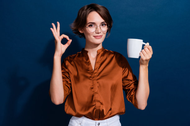 可爱的年轻女士的照片肖像拿着咖啡杯显示好符号穿着时髦的棕色衬衫孤立的深蓝色背景图片下载