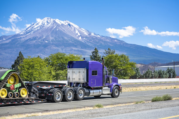 明亮的紫色强大的经典大钻机半挂车拖拉机与三个后轴运输设备的阶梯甲板半挂车运行在公路与雪山的一边图片下载