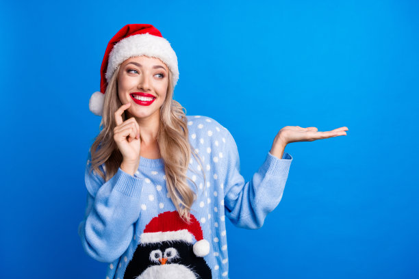 漂亮的金发女孩的照片肖像掌心看空空间穿企鹅毛衣圣诞帽孤立的蓝色背景图片下载