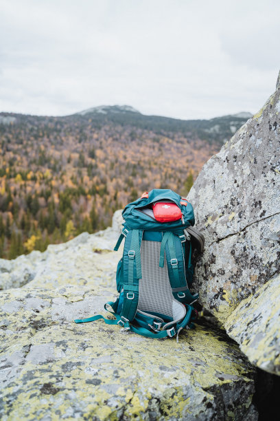 一个旅游背包矗立在岩石上，以山脉为背景，在山上徒步旅行的装备，一个红色的急救箱在包的口袋里。图片下载
