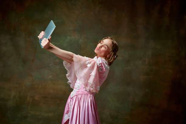 美丽的金发公主的肖像，穿着花哨的粉红色连衣裙，在平板电脑上自拍，背景是复古的纹理。典型的女孩图片下载