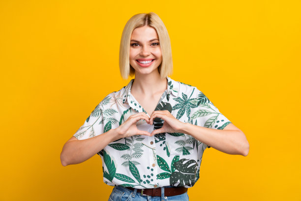 照片正面可爱的女人穿着印花衬衫微笑，露出手臂，心脏孤立的黄色背景图片下载
