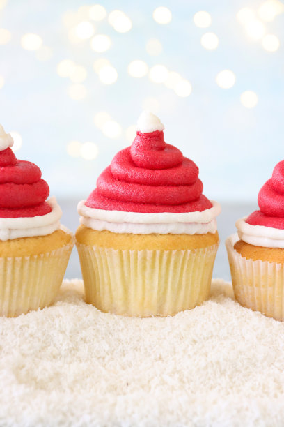 一排三个自制的、圣诞老人帽子设计的纸杯蛋糕的特写图片，红色的奶油漩涡装饰着白色的糖霜球，糖霜雪，照亮的仙女灯，淡蓝色的背景，重点在前景图片下载