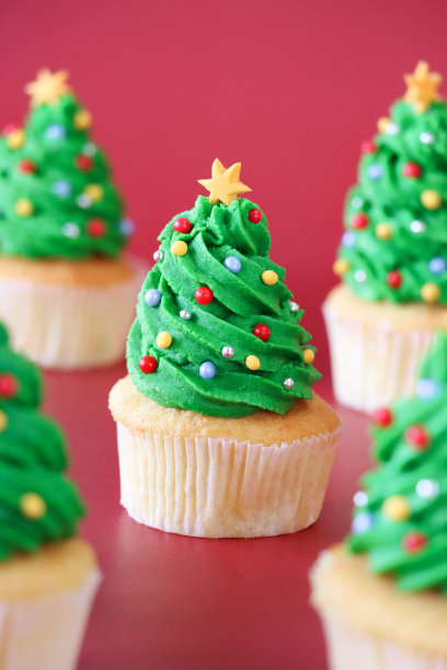 5个自制的圣诞树设计纸杯蛋糕的特写图片，绿色的奶油漩涡装饰着五颜六色的糖屑，红色背景，重点放在前景上图片下载