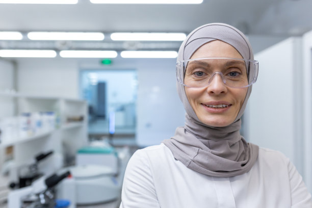 特写照片。一位年轻的穆斯林女性实验室助理、药剂师、科学家的肖像，戴着头巾和防护眼镜站在医学实验室里，微笑着看着镜头图片下载