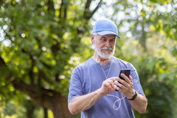 白发苍苍的老人戴着耳机用手机在公园里做运动。特写照片图片下载