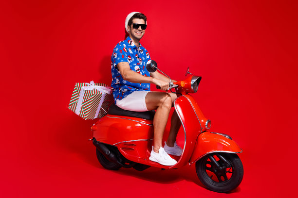 全尺寸照片时髦的年轻人驾驶摩托车送货穿着圣诞老人印花圣诞服装孤立的红色背景图片下载