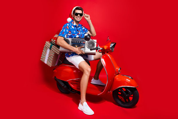 全尺寸照片时髦的年轻人驾驶滑板车送音箱穿着圣诞老人印花圣诞服装孤立的红色背景图片下载