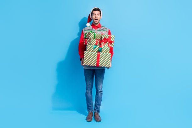 英俊的年轻人拿着堆叠礼物的全尺寸照片，震惊地穿着圣诞印花背心帽子，衣服孤立在蓝色背景上图片下载