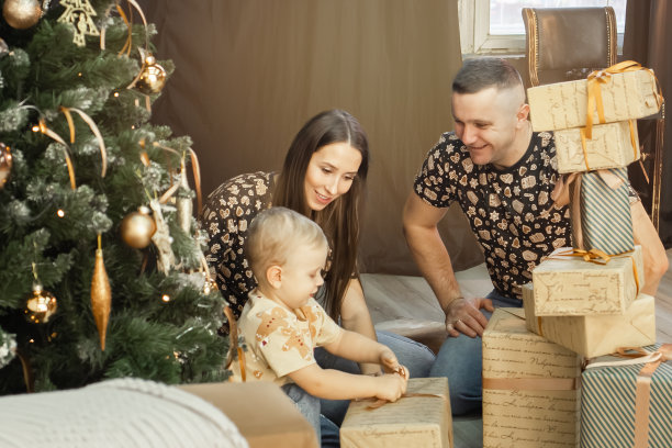 一个带着小孩的年轻家庭在圣诞树旁打开礼物的包装。一家人在客厅包装圣诞节和新年的礼物。圣诞节。新的一年图片下载