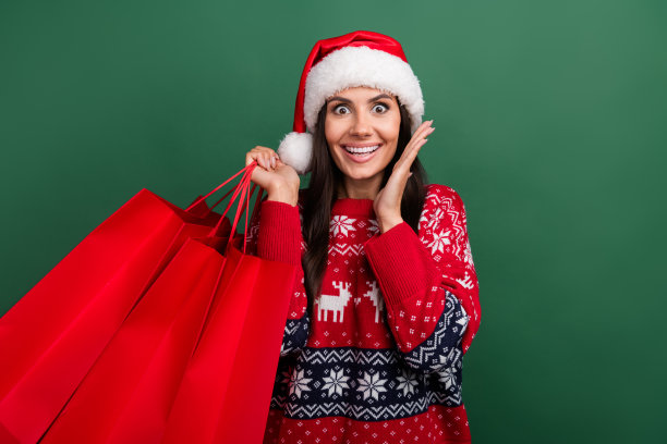 照片中，震惊的圣诞女助手穿着印花圣诞毛衣，抱着购物者，背景是孤立的绿色图片下载