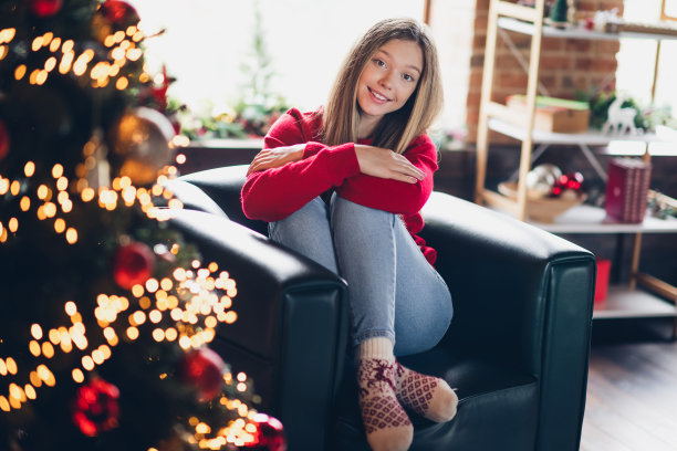 好心情的照片，漂亮的女学生，金色的发型，穿着红色的毛衣，坐在扶手椅上，抱着膝盖，在家里的圣诞树旁图片下载