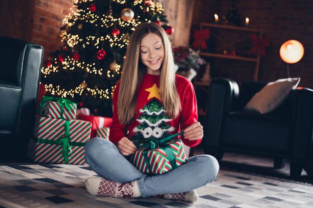积极乐观的女学生的照片，金色的发型，穿着红色的毛衣，坐在地板上打开礼盒在新年在家里室内图片下载