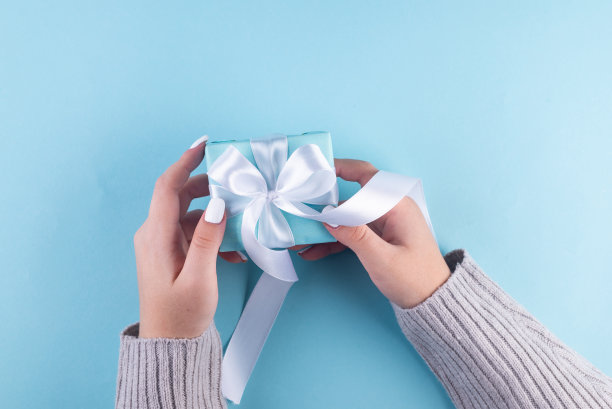 第一人称俯视图照片的女性手拿蓝色礼盒与白色丝带。柔和的蓝色背景欢迎你的话语。我的手捧着一个蓝色的礼品盒图片下载
