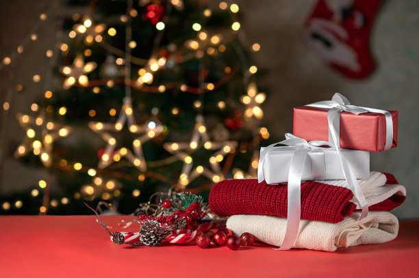 圣诞红白礼盒，针织毛衣，松树装饰，红桌子上的拐杖糖，背景上模糊闪亮的圣诞树。节日送礼，平安夜庆概念图片下载
