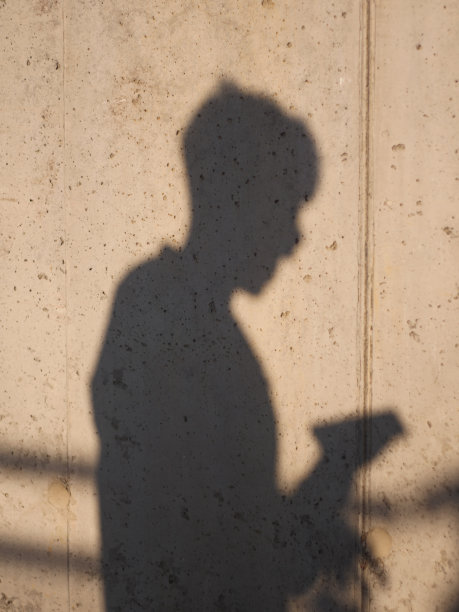 一个戴眼镜的男人看智能手机的影子剪影，混凝土工业墙上的影子，垂直概念照片图片下载