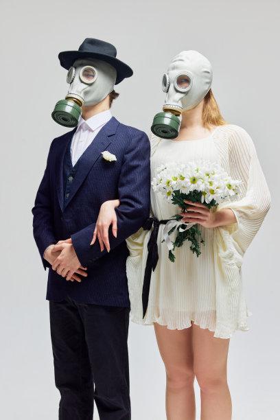 便利的婚姻，黑色西装的男人和白色礼服的女人手持鲜花，戴着防毒面具站在白色的工作室背景下。对爱和感情过敏。图片下载