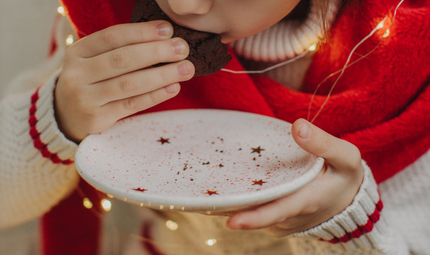 一个十几岁女孩的手拿着饼干，背景是圣诞老人的帽子和花环灯。喜庆的新年食物概念。特写照片。图片下载