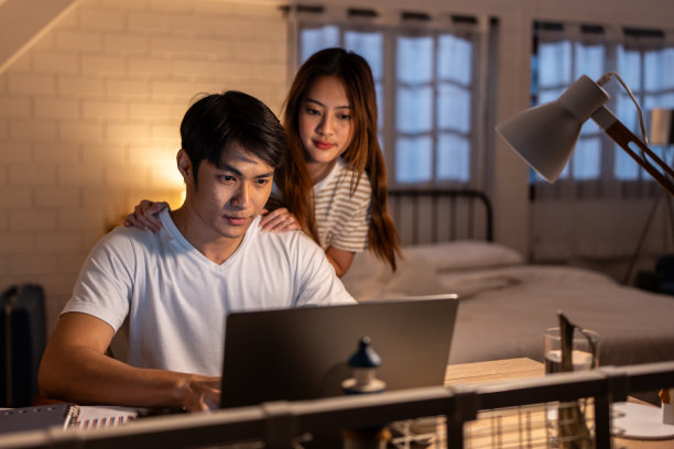 亚洲年轻人晚上在家和妻子在卧室里加班。迷人美丽的妻子穿着睡衣等着丈夫用笔记本电脑工作，在家里睡觉时上网交流。图片下载