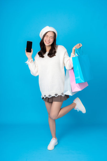 年轻亚洲女性的全身照片，网上购物，兴奋，拿着智能手机，长发，穿着白色外套。提着几个购物袋在摄影棚里拍摄蓝屏图片下载