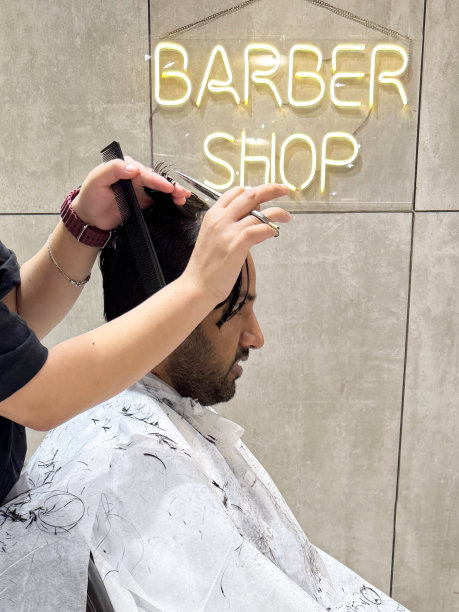 特写照片:印度男子坐在发廊的皮革扶手椅上，穿着白色的理发师斗篷，LED理发店的灯光标志，无法辨认的人在剪湿头发，塑料黑色梳子，理发师的剪刀，侧面视图，重点放在前景上图片下载