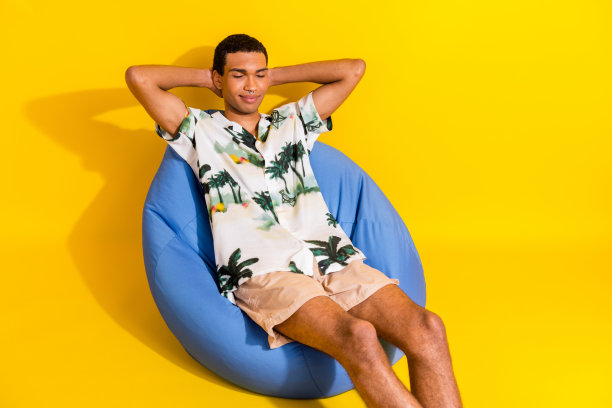 漂亮的年轻男性的照片肖像，坐在蓝色的豆袋睡觉，穿着时尚的手掌印花服装孤立在黄色的背景图片下载