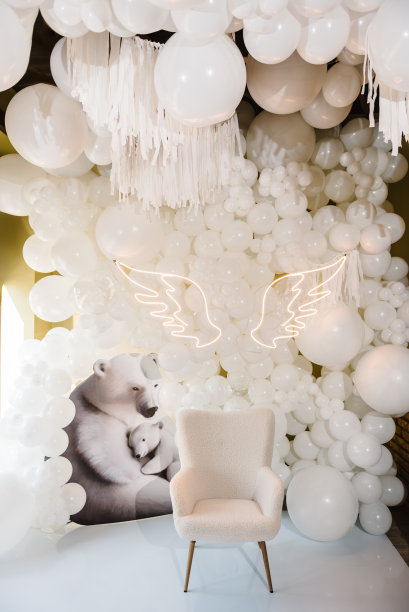 照片区以白色背景装饰空间。宝宝生日派对的接待处挂在墙上。拱门上装饰着白色气球、天使翅膀、小熊和洗礼椅。时髦的装饰。庆祝活动的概念。图片下载