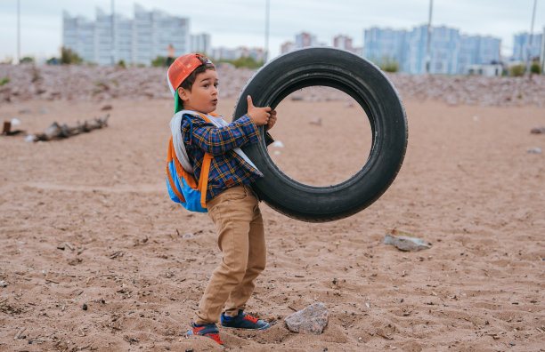 一个穿着便装的小孩在海滩上举起汽车轮胎，试图清理大自然的垃圾。生态理念，健康环境。幼儿在室外用轮子训练。童年，清新的空气留给下一代图片下载