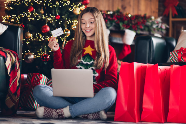 十几岁的女孩拿着信用卡的全身照片展示了如何使用笔记本电脑在网上购物时省钱的圣诞节氛围图片下载