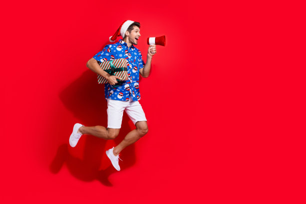 漂亮的年轻男性拿着扩音器的全身照片，穿着圣诞老人的衣服，在红色的背景上打印出圣诞服装图片下载