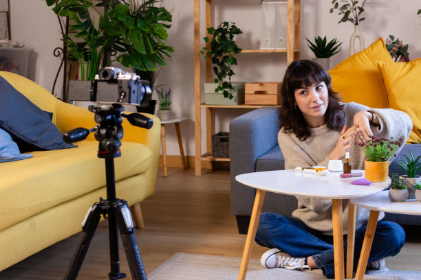 年轻的女性博主录制关于化妆品的视频博客。她坐在相机前，端着一杯咖啡。图片下载