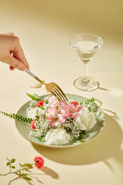 女子手里拿着一把金色的金属叉子，各种各样的鲜花陈列在一个陶瓷盘子上，旁边是一个装满水的鸡尾酒杯。杂志的艺术图片。图片下载