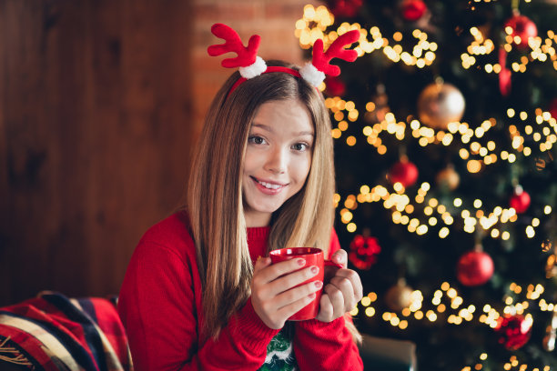 照片开朗积极美丽的女孩少年棕色头发穿红色针织毛衣坐在扶手椅拿杯子蛋酒在圣诞节的房子图片下载