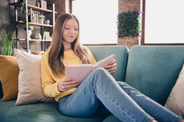 十几岁的女孩享受她在书店买的书的照片，放松地坐在客厅里，隔绝在窗户和日光的背景下图片下载