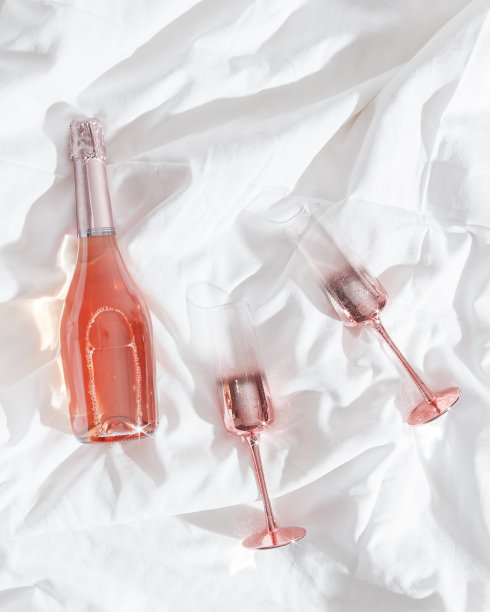 粉红色的香槟酒杯与金属光泽和一瓶玫瑰起泡酒在白色的床单。生活唯美的照片，星光滤光。情人节佳节，爱情观念，浪漫相遇。图片下载