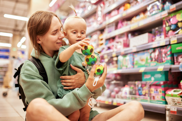 年轻迷人的女人和她可爱的孩子坐在玩具店的地板上。漂亮的妈妈和她的金发小女儿在商场购物，玩得很开心。合家周末，快乐童年。图片下载