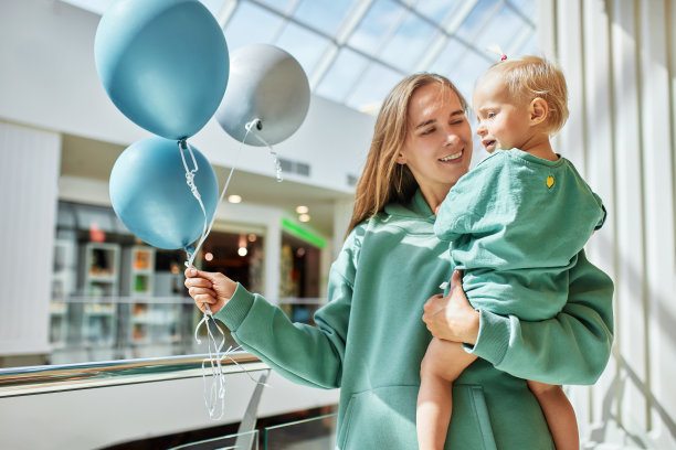 年轻迷人的母亲抱着孩子和彩色气球。快乐微笑的妈妈和她的小女儿穿着同样的绿色衣服在商场里散步，玩得很开心。家庭周末在购物中心。图片下载