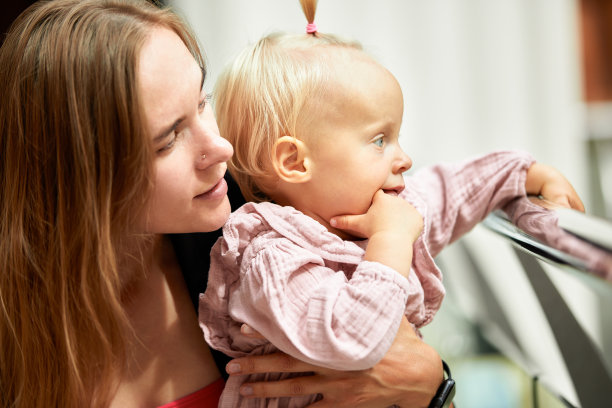 年轻漂亮的母亲的特写侧面肖像，她手里抱着一个金发小婴儿。妈妈和她可爱的女儿穿着粉红色的衣服，在购物时拥抱，放松，玩得很开心。家庭周末在购物中心。图片下载