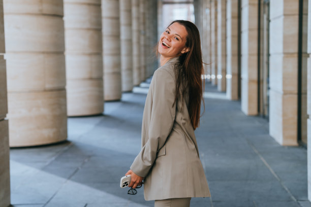 穿着西装的黑发女孩兴奋地回头看镜头，开心地微笑着享受生活，走在法院大楼的柱子之间。成功的女企业家在周五晚上、聚会时间打电话。图片下载