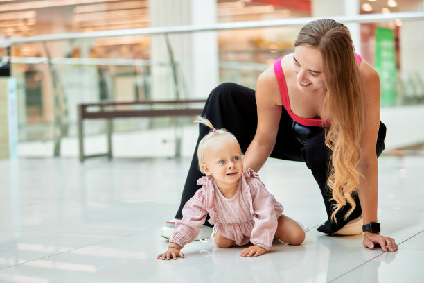 快乐的年轻妈妈看着她可爱的宝宝在公共场所、商场的地板上爬。妈妈和她的小女儿穿着粉红色的衣服在商场里散步，玩得很开心。有趣周末，快乐童年。图片下载