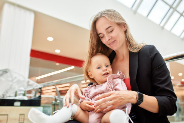 一位年轻漂亮的母亲带着一个小婴儿坐在购物中心的长椅上。穿着粉色衣服的妈妈和女儿在购物时拥抱、玩耍、放松和享受乐趣。家庭周末在购物中心。图片下载