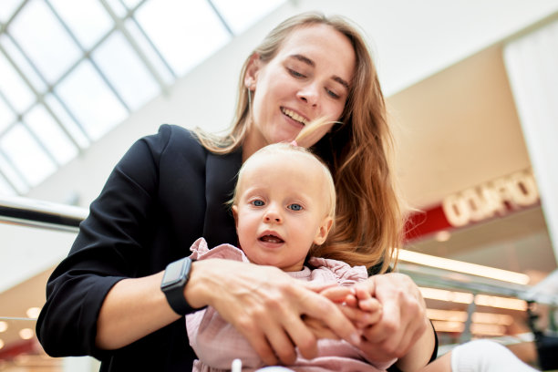 一位年轻漂亮的母亲带着一个小婴儿坐在购物中心的长椅上。穿着粉色衣服的妈妈和女儿在购物时拥抱、玩耍、放松和享受乐趣。家庭周末在购物中心。图片下载