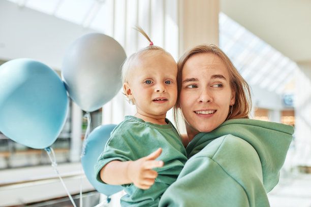 母亲的特写肖像，怀里抱着婴儿和彩色气球。年轻漂亮的妈妈带着一个穿着同样绿色衣服的小女儿在商场里散步，玩得很开心。幸福的家庭。图片下载